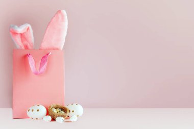 Paskalya alışverişi kompozisyonu, alışveriş çantasından tavşan kulağı, paskalya yumurtası ve pembe arka planda kuş yuvası.