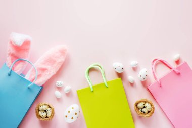 Paskalya alışverişi arka planında yeşil ve pembe alışveriş çantaları, paskalya tavşanı kulakları ve paskalya yumurtaları ve pembe arka planda kuş yuvaları var.