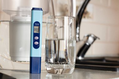 Suyun TDS 'sinin ölçümü. Elektronik pH metre ve bir bardak su. Su arıtma için filtre. Arka planda içme suyu musluğu var.. 