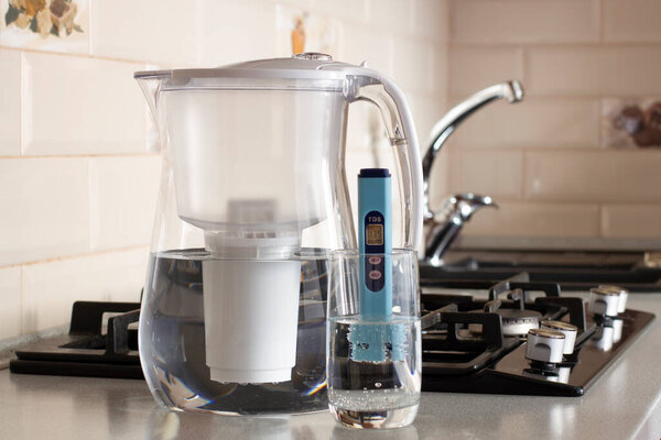 Измерение ТДС воды. Электронный pH метр в стакане воды. Фильтр для очистки воды. На заднем плане есть кран для питьевой воды. 