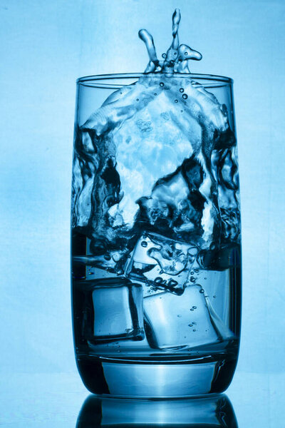 A glass of fresh clean water with splashes. A glass of water surrounded by a blue background. Drinking water with cold ice cubes