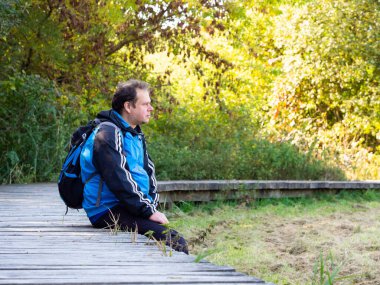 Ahşap yolda oturan bir adam, orta yaşlı bir turist. Sırtında bir sırt çantasıyla sonbaharda güneşli bir günün tadını çıkarıyor.