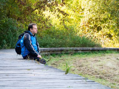 Ahşap yolda oturan bir adam, orta yaşlı bir turist. Sırtında bir sırt çantasıyla sonbaharda güneşli bir günün tadını çıkarıyor.