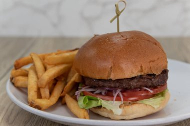Klasik bir Amerikan hamburgerinin sanatsal sunumu, tereyağlı ekmeğin üzerinde dilimlenmiş domates ve soğan ve kızarmış patatesle servis ediliyor..
