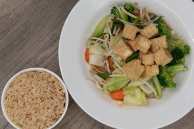Overhead view of pile of mixed vegetabels with cubes of tofu on top served with a bowl of brown rice.