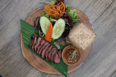 Overhead view of delicately decorated timber plate of nua yang Thai meal consisting of sliced seasoned steak, basekt of rice, and vegetable salad.