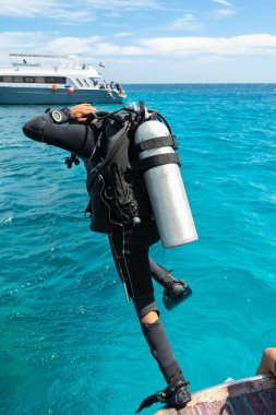 Kızıldeniz 'in sularına dalmak için tekneden tüple dalış ekipmanlarıyla atlayan bir eğitmen. Bir bacağı donmuş, diğeri de arka planda demirlemiş bir gemi olan tekneden iniyor.