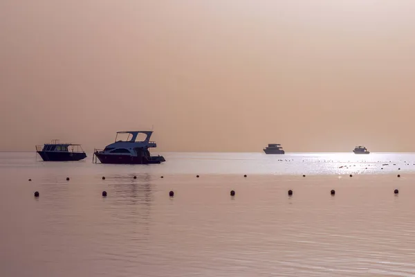 Mısır 'daki Kızıl Deniz' de şafak, Makadi Körfezi, sakin deniz, teknelerin siluetleri, ufuktaki güneşin altın diski, su yüzeyindeki güneş ışığının yansıması.