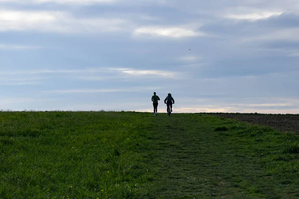 Silhouettes of a running man and a cyclist side by side against a cloudy blue sky on the horizon line