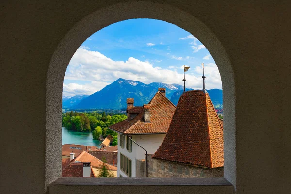 Eski Thun kasabasının kilise gözlem güvertesinin kemerli penceresinden panoramik manzara, binaların kiremitli çatıları, göl ve arka plandaki dağlar