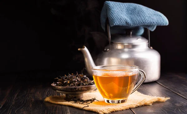 hot herbal teacup and dried tea leaves in a ceramic cup with a tea kettle placed on a black wooden table on, dark background