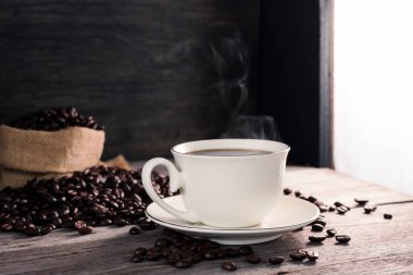 Beyaz kahve fincanında sade kahve, sıcak buhar ve koyu arka plandaki ahşap bir masaya konmuş kahve çekirdekleri. Sıcak bir atmosferde