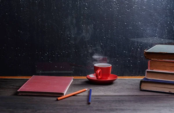 Hot tea with books on an old wooden table by the window on a rainy day.