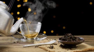 Yavaş çekim videosu, basit bir ahşap masaya yerleştirilmiş şifalı otlarla beyaz bir çay fincanına dökülen sıcak çayı gösteriyor..