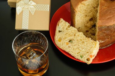 Kırmızı bir tabakta panettone dilimi, yanında bir bardak viski ve buz ve altın bir hediye kutusu..