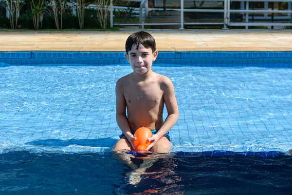 8 yaşında bir çocuk havuzun kenarında oturuyor, dudaklarını bastırıyor ve elinde bir su bombası tutuyor..