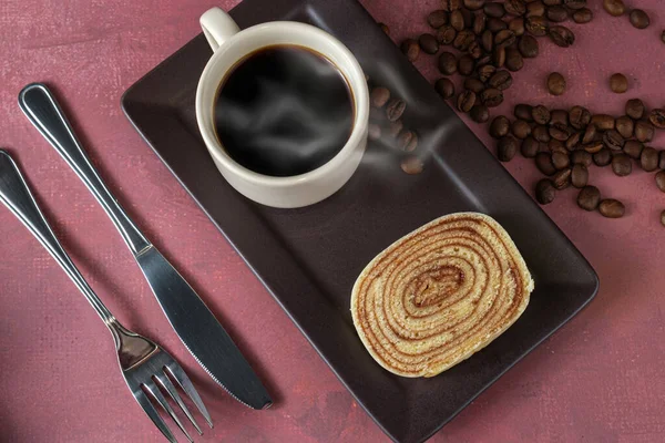 Bir dilim bolo de rolo (rulo kek) yanında çatal bıçak, fincan ve kahve çekirdekleri.