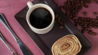 Bir dilim bolo de rolo (rulo kek) yanında çatal bıçak, fincan ve kahve çekirdekleri.