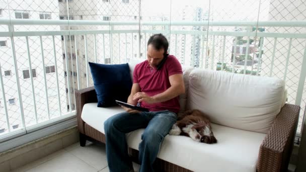 男人坐在白色的沙发上 接一个电话 在他的宠物旁边用平板电脑工作 — 图库视频影像