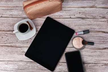 Tablet masanın üzerinde. Etrafı akıllı telefon, ruj, pres pudrası, kahve fincanı ve makyaj çantasıyla çevrili..
