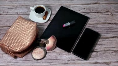 Tablet with a lipstick on it surrounded by cup of coffee smoking with cinnamon, smartphone and makeup bag.