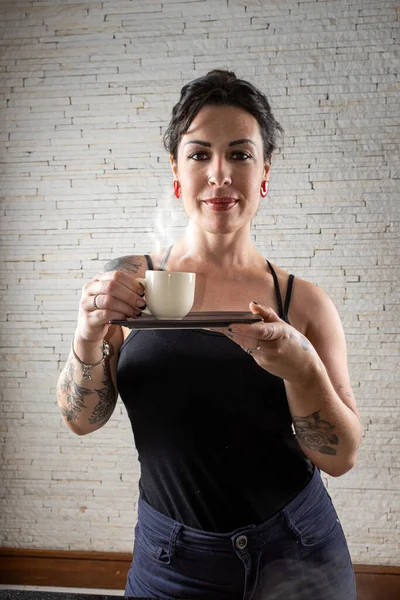 Brazilian tattooed with a slight smile, facing the camera and holding a cup of coffee.
