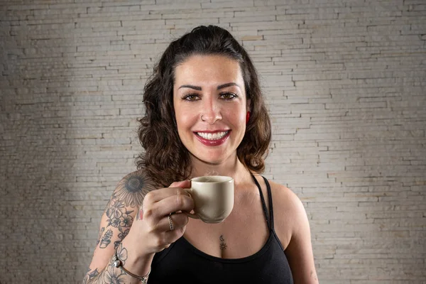 Brazilian tattooed woman with a big smile, facing the camera and holding a cup of coffee.