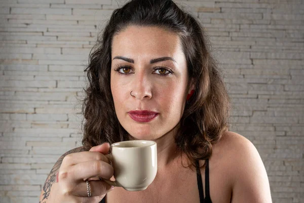 Brazilian tattooed woman facing the camera and holding a cup of coffee close to her face.