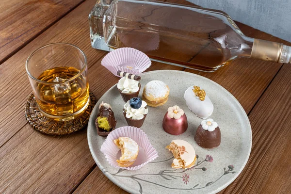 Small chocolate cup with whipped cream and blueberry, next to, walnut cameo, truffles, macaron and whiskey bottle and glass.