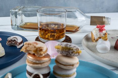 Glass of whiskey with ice surrounded by assorted wedding candies. In the blurred foreground, macarons stacked.