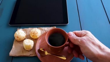 Man leaving coffee cup on table and picking up Brazilian cheese rolls next to a tablet. Blue wood background.