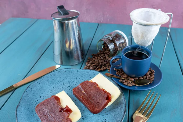 Peynirli guava tatlısı. Etrafı çatal bıçak, fincan ve kahve çekirdekleriyle çevrili. Mavi bir masada..