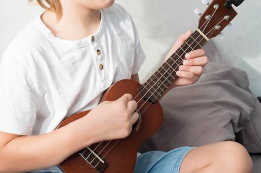 Genç çocuk evde ukulele akort ediyor. Sarı saçlı çocuk kanepede oturup akustik gitar çalıyor..