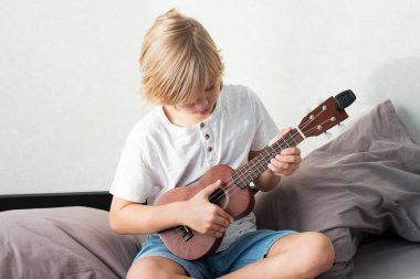 Genç çocuk evde ukulele akort ediyor. Sarı saçlı çocuk kanepede oturup akustik gitar çalıyor..