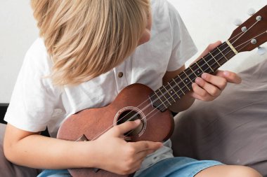 Genç çocuk evde ukulele akort ediyor. Sarı saçlı çocuk kanepede oturup akustik gitar çalıyor..
