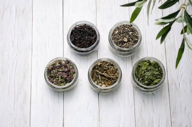 Beyaz ahşap masada cam kavanozda farklı türde bitki çayı, üst manzara. Organik, doğal kurutulmuş çay yaprakları..