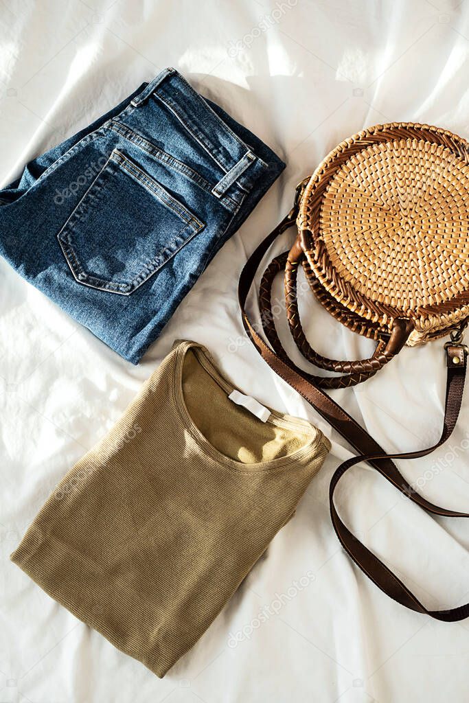 Blue jeans, trendy round rattan woven bag and khaki shirt on white bedding. Women's fashion spring or summer outfit. Stylish clothes collage, top view.