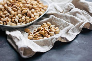 Koyu gri arka planda, açık masa örtüsünün üzerinde, kabuğunda çiğ fıstıklı bir tabak.