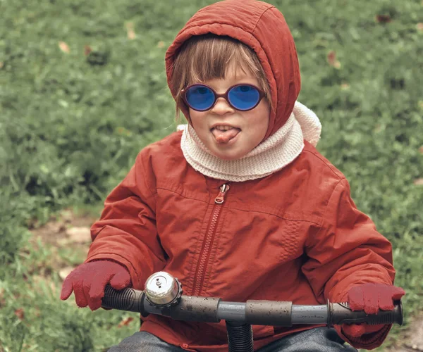 Bambina con cappuccio e occhiali blu su una piccola bici. Bambino mostra la sua lingua — Foto Stock