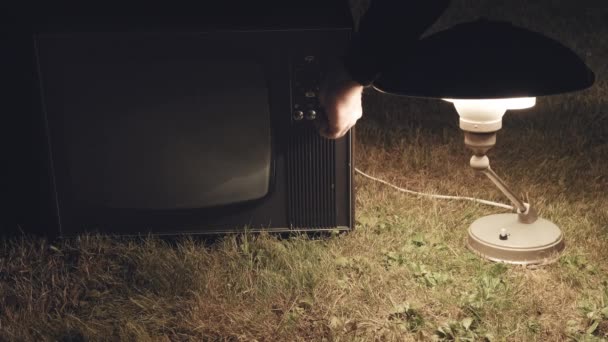 El hombre enciende un viejo televisor retro de pie en la hierba junto a una lámpara vintage — Vídeos de Stock