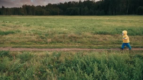 Małe, słodkie dziecko w żółtym płaszczu przeciwdeszczowym z kapturem biegnie wzdłuż drogi — Wideo stockowe