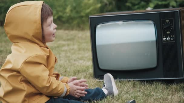 Dziecko ubrane w płaszcz przeciwdeszczowy ogląda retro TV. Dziecko klaszcze w dłonie, raduje się — Wideo stockowe