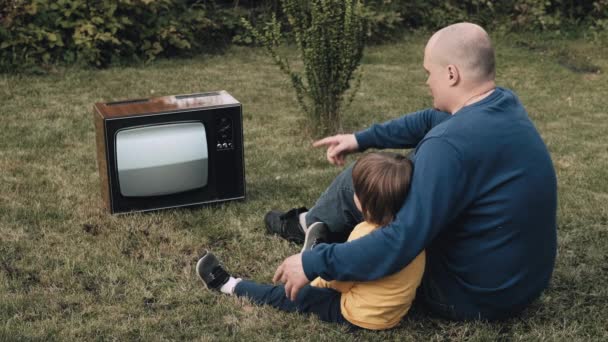 L'homme, père avec un petit enfant, est assis sur l'herbe et regarde une vieille télévision rétro — Video