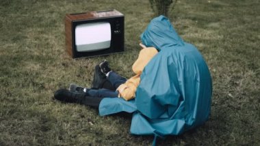 Yağmurluk giymiş küçük çocuklu bir kadın çimenlerde oturup retro televizyon izliyor.