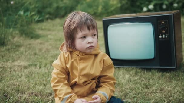 Sad child dressed in raincoat is sitting on grass. Behind him working retro TV — Stock Video