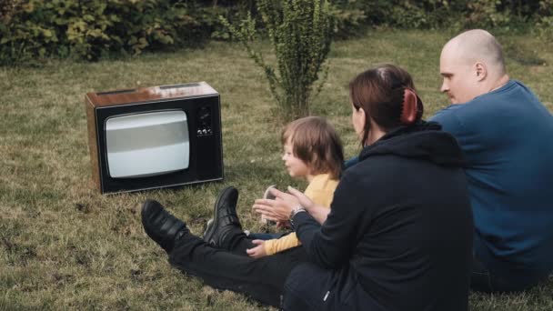 Rodina sedí na trávě a dívá se na starou retro televizi. Tleskají si. — Stock video