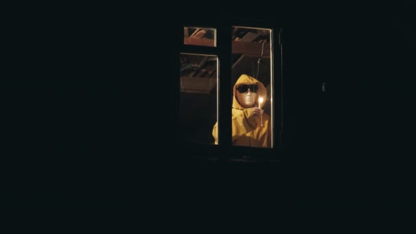 Man in raincoat and scary mask. He gives signal with candle from window — Stock Video