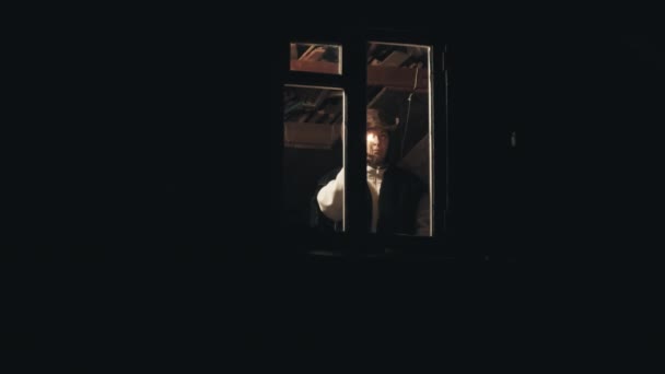 Man in cowboy hat in attic of house. He gives signal with candle from the window — Stock Video