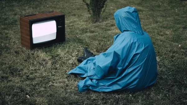 身穿蓝色雨衣头罩的男人正坐在草地上看电视 — 图库照片