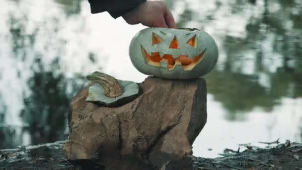 Хэллоуин. Мужчина зажигает свечи внутри тыквы на фоне реки — стоковое видео